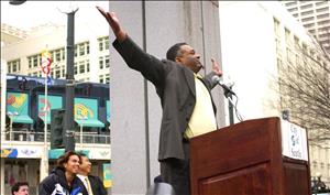 A Black man in a suit stands at a podium and throws his arms in the air while two onlookers behind him smile