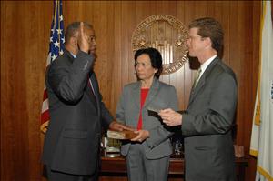 Black man in a suit raises his hand and faces a white man in a suit and places his other hand on a bible held by a white woman who is looking at him. 