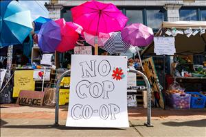 Colorful umbrellas, signs, and crates of fruit behind a sign that says No Cop Co Op