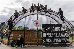A group of white people wearing black clothing and face masks are sitting on top of a metal batters cage above signs that say Aid Station and Why Do We Have to Keep Telling You Black Lives Matter