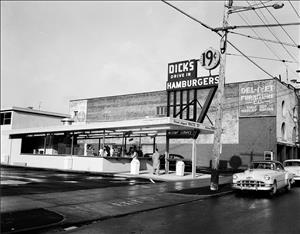 A one story drive-in restaurant with two people at the take out window and mid-century cars parked along the street in front.