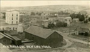 Elevated view, train tracks and warehouse in front, downtown commercial buildings behind, scattered homes on slope rising at rear