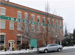Three story brick building with many arched windows and a green sign painted on the front that says Broadview Dairy. In front of the building there is a snow on the sidewalk and one gray car parked. 