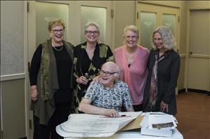 Four elderly white women stand behind a seated white man in glasses who sits at a table covered with documents and sketches. They are in a room that is all beige and brown. 