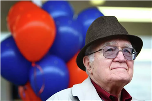 Head and shoulders shot of white-haired man in fedora and glasses in front of red and blue balloons  