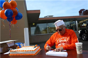 White-haired man wearing chef's hat and orange T-shirt reading "Celebrating Dick's" sitting at table outside restaurant, signing papers, next to decorated cake and red and white balloons   