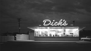 Black and white photo of one-story restaurant stand at night, empty interior and neon "Dick's" sign on roof lit and glowing