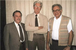 Three gray-haired men, in jacket and tie, sweater and tie, and white shirt and vest, smiling at camera
