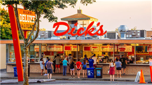 People standing in front of drive-in, with menus and servers visible in windows beyond them and neon "Dick's" sign on roof