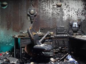 Photo of the control room of The Dutchman rehearsing the recording studio after fire of January 9, 2009. Everything is scorched black, melted computer monitors sit on a charred desk, beneath which are various pieces of destroyed electrical equipment. A burned guitar is visible behind a ruined chair, and the decorative clock on the wall is blackened by soot.