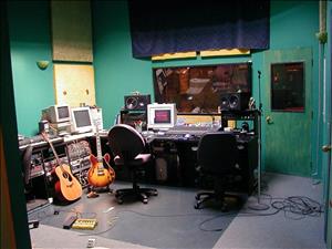 Control room of The Dutchman rehearsal and recording studio in 2000. A small room with green walls, with two guitars, three computer monitors, and miscellaneous recording equipment. In front of the control panel is a window that looks into the rehearsal and recording space.