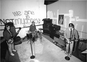 A three piece band, The Statics, in the recording room of The Dutchman rehearsing and recording studio. Written on the back wall is "We are NOT Mudhoney," a reference to another local band. From left to right are the bass player, the drummer, and the guitarist.