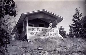 Black and white photo looking up rocky, grassy slope to a small building with woman in suit seated on front porch railing above sign reading R. B. Exton Real Estate