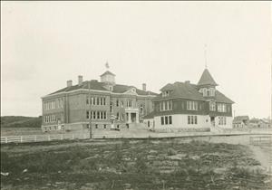 Large white-trimmed brick building at center, smaller wood frame building in front on the right, very small third building barely visible between the others