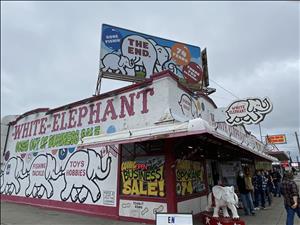 People lined up outside a one story white building with the words White Elephant and Sale painted on the side and a billboard on the roof that says The End