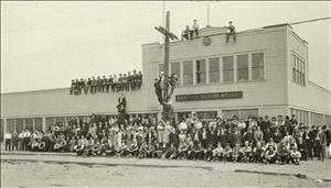 Kilbourne and Clark Manufacturing Company building in SoDo, a mostly one story wood-frame structure with a second story added above the entryway to the right. Crowding the street, lining a portion of the roof, and hanging from a utility pole are the company's employees. The photo was taken in June 1922