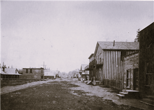View of dirt street with group of people standing near wood buildings on right side