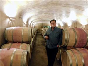 A white man with a mustache and glasses holds a glass of red wine and leans against a barrel in a cavernous room full of barrels