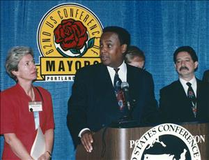 Seattle Mayor Norm Rice at speakers podium with wall placard behind reading "62nd US Conference of Mayors, Portland," with three unidentified people