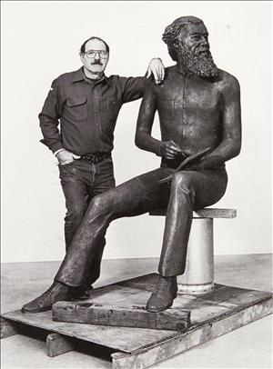Levine posing with glasses, work shirt and jeans next to a statue of John Muir, sitting on a pedestal that is fixed to a wooden palette