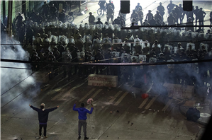 Birds eye view of two people facing several rows of riot police. One person is holding their arms out to the side. The other holds flowers. There are clouds of smoke and gas in the air. It is nightime. 