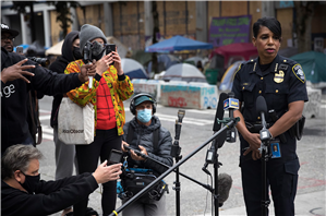 A Black woman in an police uniform addresses a crowd of people holding cameras and microhones. Behind her is graffiti and concrete barricades