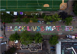 Birds eye view of a street with the words Black Lives Matter painted in bright colors and patterns on the surface of the street. On one side of the street, a sports field, and on the other the tops of buildings. 