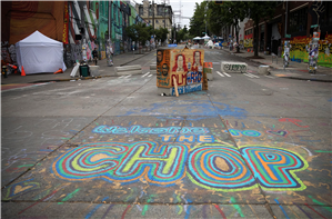 Looking up a street that is covered in graffiti, tents, and street barricades. The word CHOP is written on the street in brightly colored chalk or paint. 