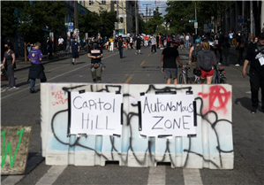 Pedestrians in summer clothes walk and push bikes down the middle of a street that is blockaded with concrete blocks, in the foreground, with handmade signs that say Capitol Hill Autonomous Zone