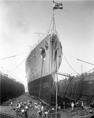 A large ship flying a german flag sits on rails and is chained to walls on either side. Men below it are working. 
