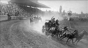 Two stage coaches with four horses and at least three men each racing across a dirt track as a crowd looks on in stadium seating