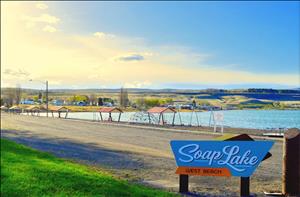 Color image of a blue and orange sign that says Soap Lake West Beach with a playground, a lake, and some houses in the background