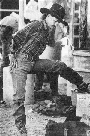 Leslie with his leg up stretching in cowboy hat, plaid flannel, jeans and cowboy boots outside with other cowboys in the background