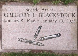 A flat tombstone in the grass with the name of the deceased and engraved images of crayons. The text on the stone says Seattle Artist Gregory L Blackstock