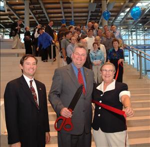 Three people smile for the camera, one holding giant red scissors and the other holding a red ribbon that continues up the stairs behind them where a crowd looks on. 