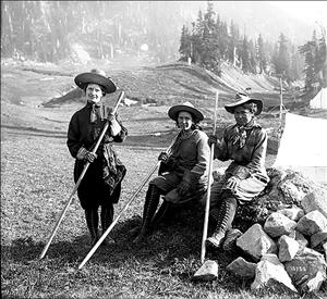 Three woman in early 20th century hiking clothes, boots, and sun hats, all hold hiking sticks in a meadow with tents and forest behind them