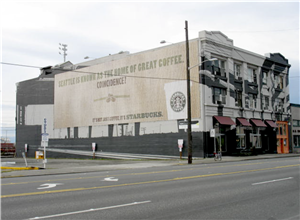 A three story commercial building on a four lane street next to a parking lot. The side of the building is a painted advertisement for starbucks that says seattle is known as the home of great coffee coincidence. There are red awnings over the front windows and a large orange front door. 