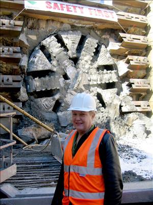 A woman in a white hard hat and safety orange vest stands in front of a large round piece of equipment with the sign Safety First above. 