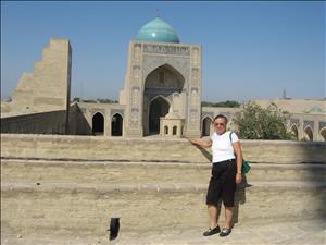 A woman stands in front of a sand colored mosque with elaborate tile work and a blue minaret. 