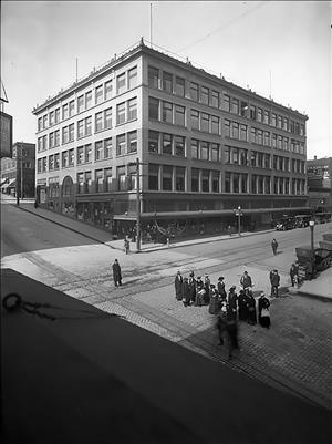 A five story stone building on the corner of an intersection with several people crossing the street. 