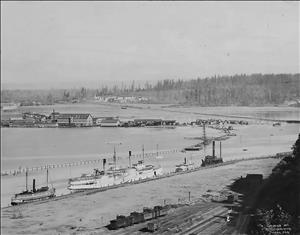 Ships, railroad cars, and factories along the water's edge in a bay surrounded by forested hillsides.