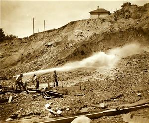 Three men work next to a large hose spraying a large plume of water over a muddy embankment. There is a wooden house on the bluff above them. 