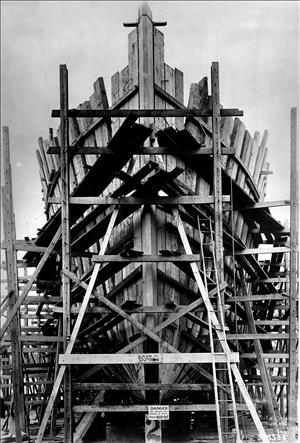 The bow of a wooden ship under construction with thick wood frames surrounded by wooden scaffolding. 