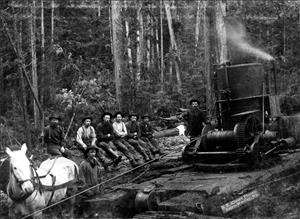 Men in work clothes, hats, and boots sit on a large log in the forest. One man operates machinery and another stands beside a white horse. Image is black and white. 