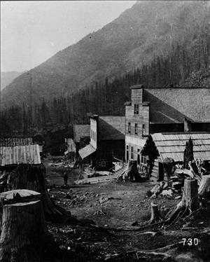 Old wooden buildings along a dirt road with tree stumps in it. A forested hillside in the background. 