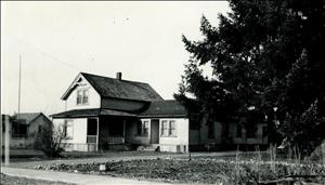 A two story wooden house with a covered porch and a large evergreen tree in front. 