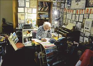 A white man with white hair rests his head on one hand and leans over a desk covered in colorful sketches. The room is very cluttered with books art musical instruments and other ephemera
