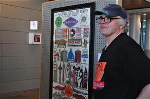 A white man in a cap and glasses smiles and stands in front of a large print covered in colorful sketches