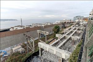 Concrete, rebar, rubble, and pillars in the foreground. Rooftops along a large body of water in the background. 