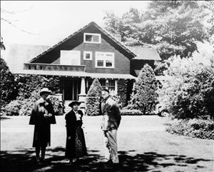 Three people stand in front of a large house surrounded by shrubs and trees. 